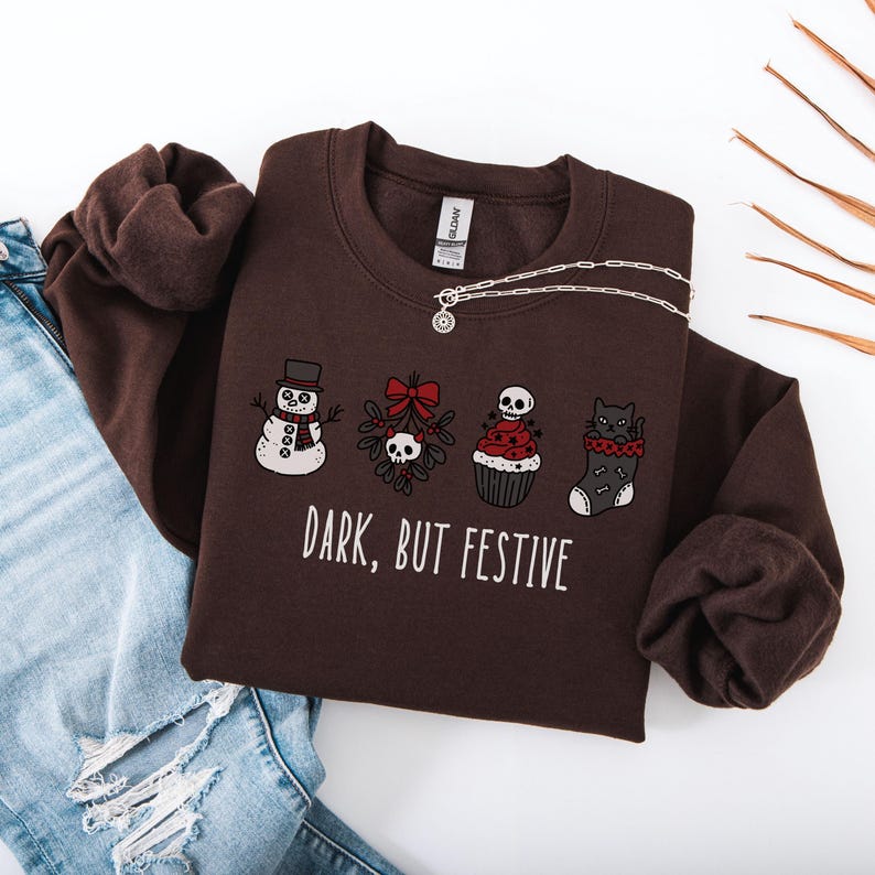 Op de afbeelding: Een donkerbruine sweater met ronde hals en een grafische print van een sneeuwpop, maretak met een schedel, een cupcake met een schedel als topping en een kat in een kerstsok. De tekst "DARK, BUT FESTIVE" staat in witte letters.