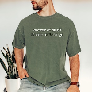 Op de afbeelding: Olijfgroen t-shirt met de witte tekst "knower of stuff fixer of things". Het shirt heeft een relaxte pasvorm en korte mouwen. De persoon draagt een bruin horloge.