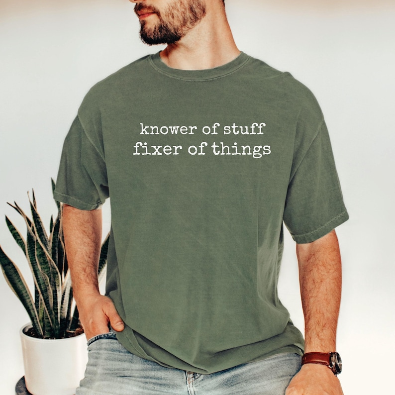 Op de afbeelding: Olijfgroen t-shirt met de witte tekst "knower of stuff fixer of things". Het shirt heeft een relaxte pasvorm en korte mouwen. De persoon draagt een bruin horloge.