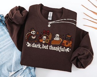 May include: A dark brown crewneck sweatshirt featuring a graphic print of a skull-headed turkey, a cornucopia with a skull, a slice of pie with a skull, and a black cat in a pumpkin. The text reads "dark, but thankful" in white lettering.