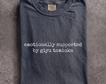 May include: A dark gray short-sleeved t-shirt with a crew neckline. The shirt features white text printed on the chest that reads, "emotionally supported by giyu tomioka." A tan brand label is visible inside the collar of the garment.