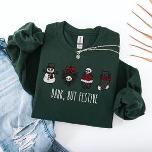 Op de afbeelding: Een donkergroen sweatshirt met lange mouwen en een grafische print van een sneeuwpop, maretak met een schedel, een cupcake met een schedel als topping en een zwarte kat in een kerstsok. De tekst "DARK, BUT FESTIVE" staat in witte hoofdletters.