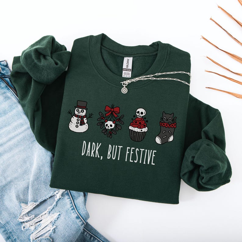Op de afbeelding: Een donkergroen sweatshirt met lange mouwen en een grafische print van een sneeuwpop, maretak met een schedel, een cupcake met een schedel als topping en een zwarte kat in een kerstsok. De tekst "DARK, BUT FESTIVE" staat in witte hoofdletters.