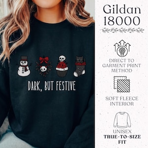 Op de afbeelding: Een zwart sweatshirt met ronde hals en een grafische print van een sneeuwpop, een schedel met een rode strik, een cupcake met een schedel en een zwarte kat in een kerstsok. De tekst luidt "DARK, BUT FESTIVE" met extra productinformatie.
