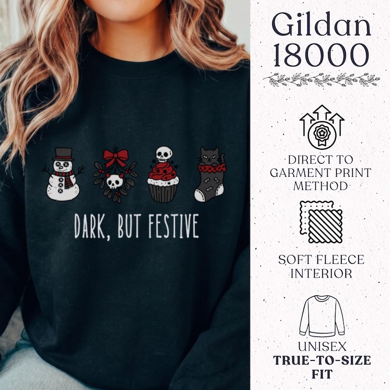 Op de afbeelding: Een zwart sweatshirt met ronde hals en een grafische print van een sneeuwpop, een schedel met een rode strik, een cupcake met een schedel en een zwarte kat in een kerstsok. De tekst luidt "DARK, BUT FESTIVE" met extra productinformatie.