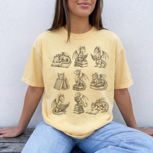 K&ouml;nnte beinhalten: Ein blassgelbes Kurzarm-T-Shirt mit einem dunkelbraunen Grafikdruck von neun kleinen Drachen in verschiedenen Posen, die mit B&uuml;chern interagieren. Die Drachen sind beim Lesen, Sitzen auf oder Schlafen neben Stapeln von offenen und geschlossenen B&uuml;chern zu sehen.