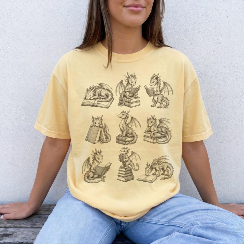 K&ouml;nnte beinhalten: Ein blassgelbes Kurzarm-T-Shirt mit einem dunkelbraunen Grafikdruck von neun kleinen Drachen in verschiedenen Posen, die mit B&uuml;chern interagieren. Die Drachen sind beim Lesen, Sitzen auf oder Schlafen neben Stapeln von offenen und geschlossenen B&uuml;chern zu sehen.