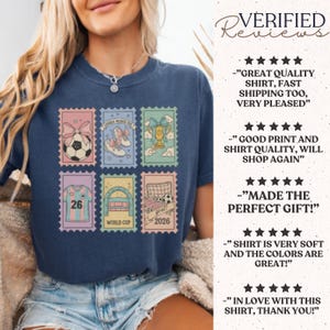 May include: Navy blue t-shirt featuring six postage stamp designs with soccer-themed graphics. The stamps include a soccer ball, shoes, a trophy, a jersey with the number 26, a stadium, and a soccer goal with the text "goal 2026".