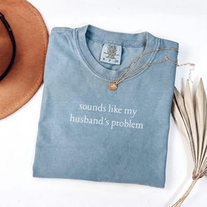 May include: A blue short-sleeved t-shirt with a crew neckline featuring the white printed text "sounds like my husband's problem" on the chest. The shirt has a Comfort Colors brand label at the inner neck and is displayed with a gold-toned necklace.