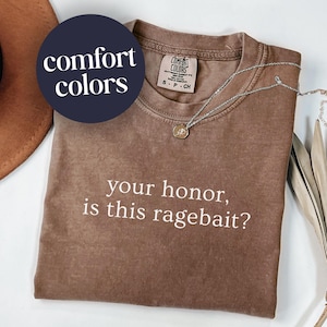 May include: A brown Comfort Colors t-shirt with the phrase "your honor, is this ragebait?" in white text. A silver necklace with a pendant is draped on the shirt.