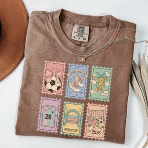 May include: A brown Comfort Colors t-shirt featuring six colorful postage stamp designs with soccer-themed graphics. The stamps include a soccer ball, cleats, a trophy, a jersey with the number 26, and the text "WORLD CUP 2026". A silver necklace is draped across the shirt.