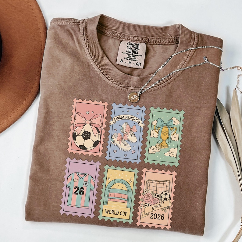 May include: A brown Comfort Colors t-shirt featuring six colorful postage stamp designs with soccer-themed graphics. The stamps include a soccer ball, cleats, a trophy, a jersey with the number 26, and the text "WORLD CUP 2026". A silver necklace is draped across the shirt.