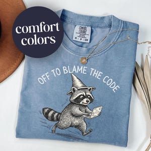 May include: A light blue Comfort Colors t-shirt with the text "OFF TO BLAME THE CODE" and a cartoon raccoon wearing a wizard hat and holding a laptop. A gold necklace is draped on the shirt.