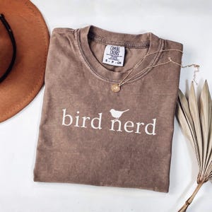 Pu&ograve; includere: Una t-shirt marrone a maniche corte con scollo rotondo e una stampa grafica bianca che recita bird nerd con una piccola icona di uccello. La maglietta presenta un'etichetta del marchio Comfort Colors sul collo ed &egrave; in tessuto morbido e slavato.