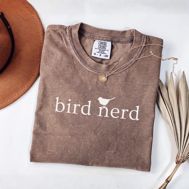 Pu&ograve; includere: Una t-shirt marrone a maniche corte con scollo rotondo e una stampa grafica bianca che recita bird nerd con una piccola icona di uccello. La maglietta presenta un'etichetta del marchio Comfort Colors sul collo ed &egrave; in tessuto morbido e slavato.