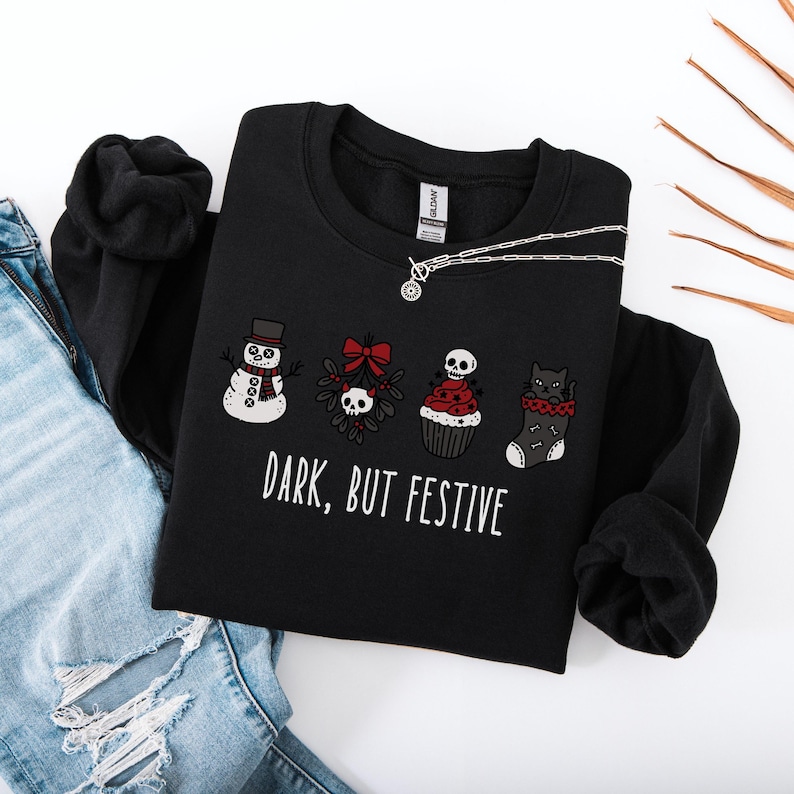 Op de afbeelding: Een zwart sweatshirt met lange mouwen en een afbeelding van een sneeuwpop, maretak met een schedel, een cupcake met een schedel en een kat in een kerstsok. De tekst "DARK, BUT FESTIVE" staat in witte letters. Gecombineerd met een blauwe spijkerbroek.