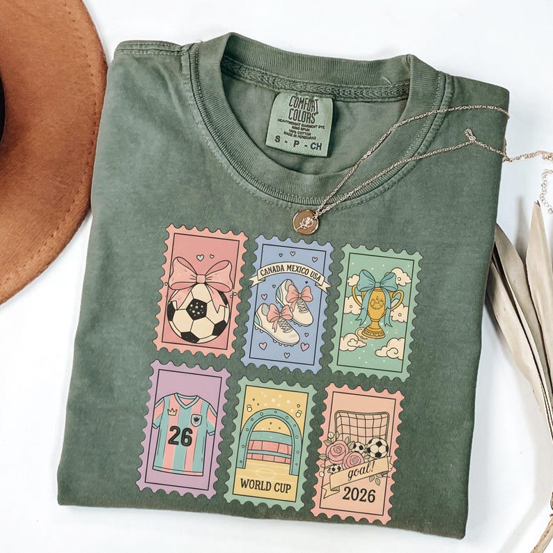 May include: Olive green t-shirt with a soccer-themed stamp design. The stamps feature soccer balls, shoes, a trophy, and the text "CANADA MEXICO USA" and "WORLD CUP 2026". A silver necklace with a pendant is also visible.