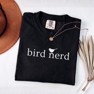 Pu&ograve; includere: Una t-shirt nera a maniche corte con una stampa grafica bianca che recita bird nerd, con una piccola icona di uccello sopra la lettera r. La maglietta ha un girocollo e un'etichetta del marchio Comfort Colors, taglia S-P-CH.