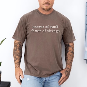 Op de afbeelding: Een bruin t-shirt met de tekst "knower of stuff fixer of things" in het wit. Het shirt heeft korte mouwen en een ronde hals. Het shirt is gemaakt van een zacht materiaal.