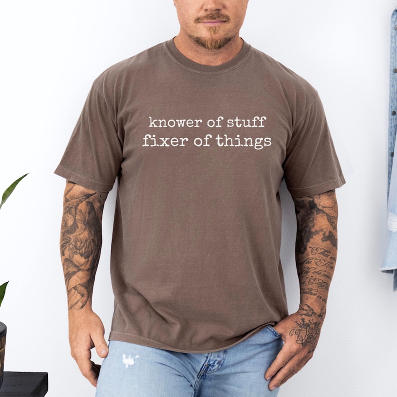 Op de afbeelding: Een bruin t-shirt met de tekst "knower of stuff fixer of things" in het wit. Het shirt heeft korte mouwen en een ronde hals. Het shirt is gemaakt van een zacht materiaal.