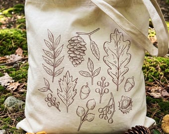 May include: A cream-colored canvas tote bag featuring a brown line-art illustration of various forest elements, including a pinecone, an oak leaf, acorns, berries, and assorted foliage. The bag has two fabric handles and a rectangular shape.