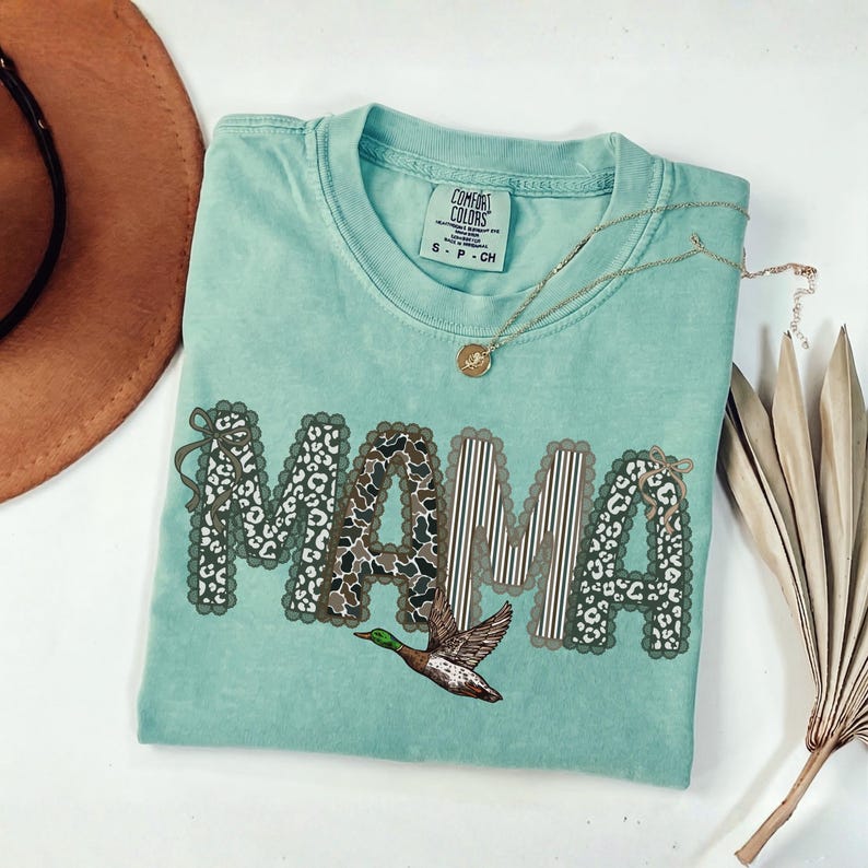 Peut inclure: Un t-shirt vert clair avec un col rond et une &eacute;tiquette de marque Comfort Colors. Le devant pr&eacute;sente le mot MAMA en grandes lettres aux bords festonn&eacute;s, d&eacute;cor&eacute; de motifs l&eacute;opard et ray&eacute;s, avec un graphique de canard colvert en vol.