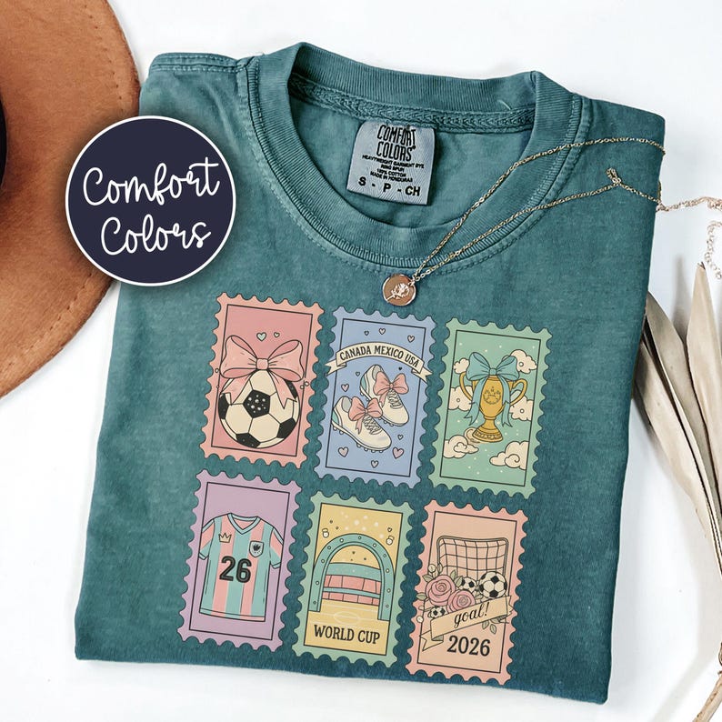 May include: Teal Comfort Colors t-shirt featuring six colorful postage stamp designs with soccer-themed illustrations. The stamps include a soccer ball, soccer shoes, a trophy, a jersey with the number 26, and the words "World Cup" and "goal! 2026".