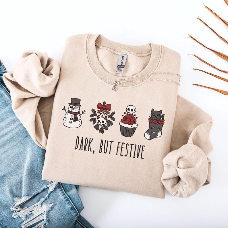 Op de afbeelding: Een beige sweater met ronde hals en een grafische print van een sneeuwpop, maretak met een schedel, een cupcake met een schedel erop en een kat in een kerstsok. De tekst "DARK, BUT FESTIVE" staat in zwarte letters onder de illustraties.