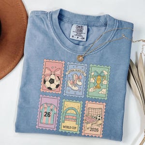 May include: A light blue Comfort Colors t-shirt featuring six postage stamp designs with soccer-themed illustrations. The stamps include a soccer ball, soccer shoes, a trophy, a jersey with the number 26, a soccer goal, and the text "goal! 2026". A gold necklace is draped across the shirt.