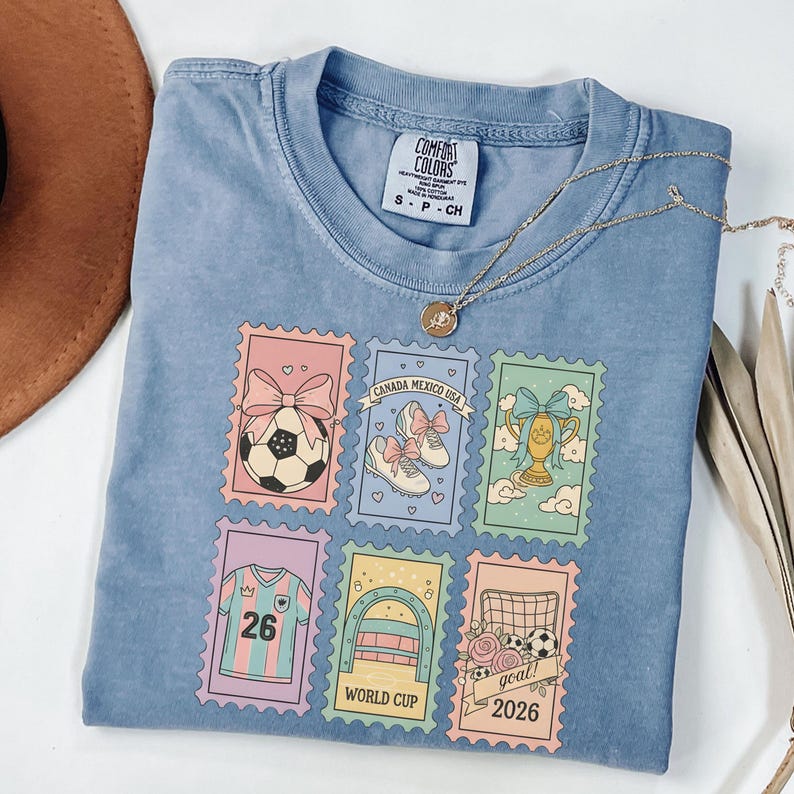 May include: A light blue Comfort Colors t-shirt featuring six postage stamp designs with soccer-themed illustrations. The stamps include a soccer ball, soccer shoes, a trophy, a jersey with the number 26, a soccer goal, and the text "goal! 2026". A gold necklace is draped across the shirt.