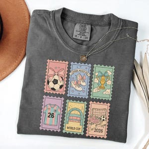 May include: Dark grey t-shirt with a necklace. The shirt features colorful postage stamp designs with soccer-themed graphics, including a soccer ball, shoes, a trophy, a jersey, a goal, and the text "WORLD CUP 2026".
