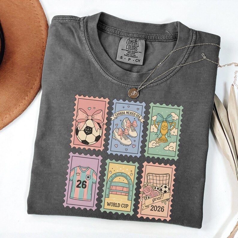 May include: Dark grey t-shirt with a necklace. The shirt features colorful postage stamp designs with soccer-themed graphics, including a soccer ball, shoes, a trophy, a jersey, a goal, and the text "WORLD CUP 2026".