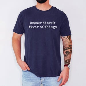 Op de afbeelding: Marineblauw t-shirt met de witte tekst "knower of stuff fixer of things". Het shirt is effen van kleur en heeft korte mouwen. De persoon draagt ook een blauwe spijkerbroek.