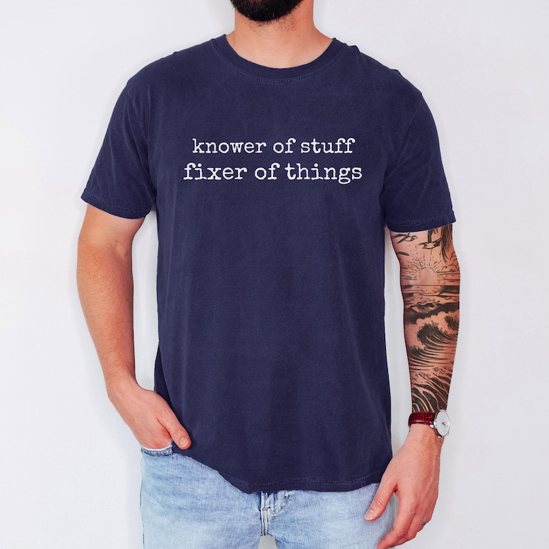 Op de afbeelding: Marineblauw t-shirt met de witte tekst "knower of stuff fixer of things". Het shirt is effen van kleur en heeft korte mouwen. De persoon draagt ook een blauwe spijkerbroek.