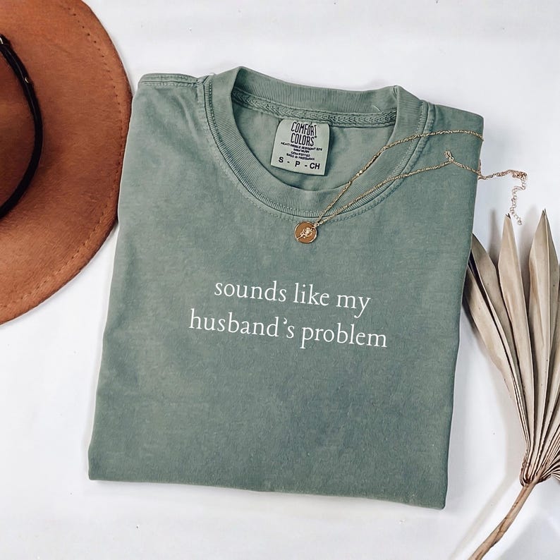 May include: A sage green short-sleeve t-shirt with a crew neckline and the white text "sounds like my husband's problem" printed on the chest. The shirt features a Comfort Colors brand label at the inner neck and is paired with a gold pendant necklace.