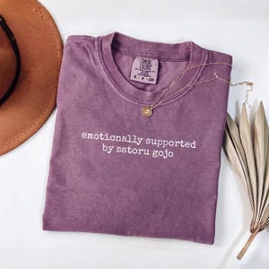 May include: A mauve short-sleeved t-shirt with a crew neckline featuring the white text "emotionally supported by satoru gojo" printed on the chest. The shirt has a Comfort Colors brand label inside the collar indicating a size small.