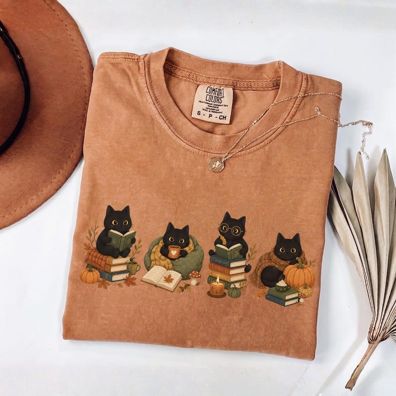 May include: A rust-colored t-shirt with a Comfort Colors label at the neck. The graphic features four black cats in autumn settings, reading books, drinking from mugs, and sitting near pumpkins, mushrooms, and fall leaves. The shirt has a crew neckline.