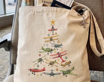 May include: A cream-colored canvas tote bag featuring a graphic print of various vintage-style airplanes arranged in the shape of a Christmas tree, accented with small stars and ornaments. A silver airplane keychain is attached to the strap.