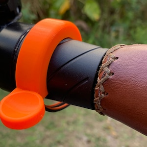 May include: Close-up of an orange bicycle bell attached to a black handlebar. The bell has a circular design with a small lever. The handlebar grip is brown leather with a stitched pattern.
