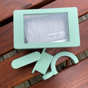 May include: A mint green box with a clear window showing a white and gray patterned fabric with the text "bubu kiki by Cin." The box is open and there are two mint green plastic pieces next to it.