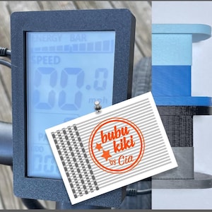 May include: A black electric bike display with a digital screen showing the speed in miles per hour. The screen also shows a battery bar. The display is mounted on the handlebars of the bike. A white card with a red circle and the text "bubu kiki by Cia" is attached to the display.
