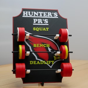 May include: A black sign with the text "HUNTER'S PR'S" and the words "SQUAT", "BENCH", and "DEADLIFT" written in white. The sign has a red cardinal logo and three sets of weight plates with black bars.