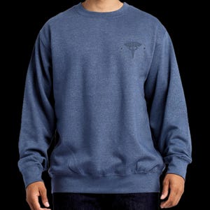 May include: A heathered blue crewneck sweatshirt featuring a circular emblem on the upper left chest. The emblem contains a caduceus symbol at the center, surrounded by text that reads "EMT" and "PARAMEDIC" along the circular border.