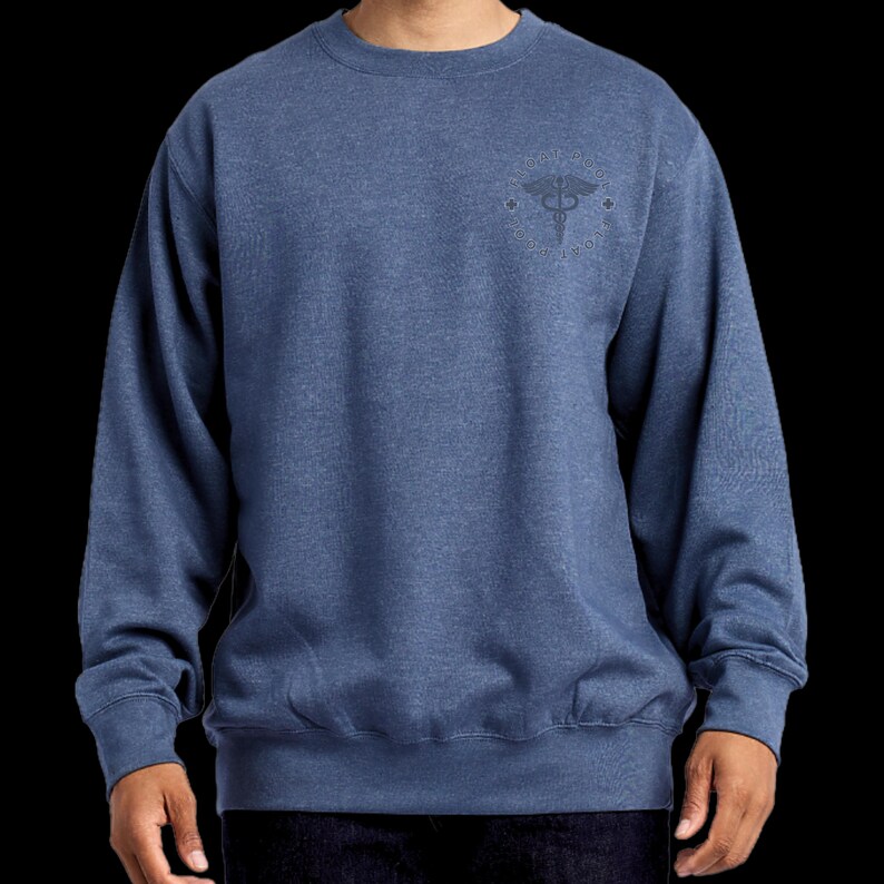 May include: A heathered blue crewneck sweatshirt featuring a circular emblem on the upper left chest. The emblem contains a caduceus symbol at the center, surrounded by text that reads "EMT" and "PARAMEDIC" along the circular border.