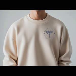May include: A beige crewneck sweatshirt featuring a circular navy blue graphic on the left chest. The design includes a caduceus symbol in the center, surrounded by the text "FLOAT POOL" and two small cross icons on the left and right sides.