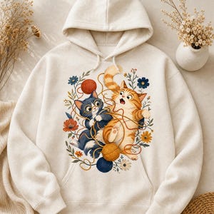Puede incluir: Una sudadera con capucha de color crema que presenta un gráfico central de un gato azul oscuro con gafas y un gato atigrado naranja jugando con ovillos de lana de colores, rodeados de pequeñas flores y follaje en tonos azules, naranjas y verdes.