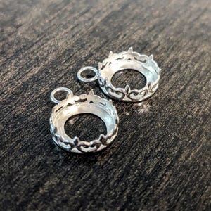 May include: Two silver-toned pendant settings with ornate, decorative edges. The settings are round and have a hole at the top for attaching a chain or cord.