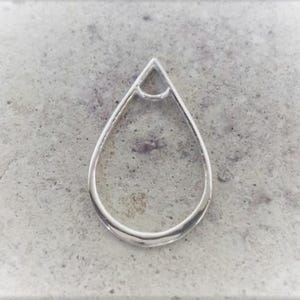 May include: A silver-colored, teardrop-shaped pendant with a small triangle at the top. The pendant is set against a textured, gray background. The design is simple and elegant.