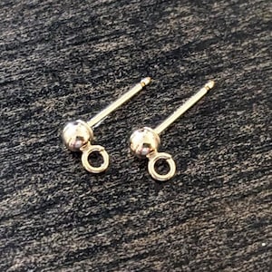 May include: A pair of gold-colored earring posts with a ball-shaped stud and a small loop. The posts are on a dark, textured surface.