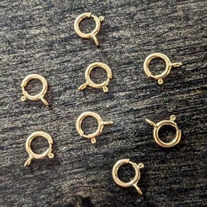 May include: A collection of seven gold-toned circular spring ring clasps, each featuring a small attached loop for jewelry assembly. These metallic findings are designed with a spring-loaded mechanism for opening and closing, commonly used as fasteners for necklaces or bracelets.